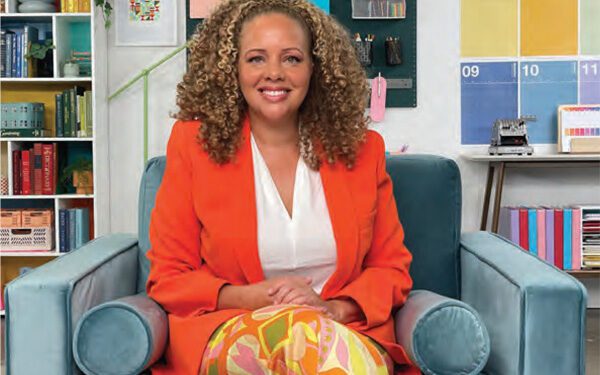 woman seated in chair with bright orange blazer and stacks of books behind her